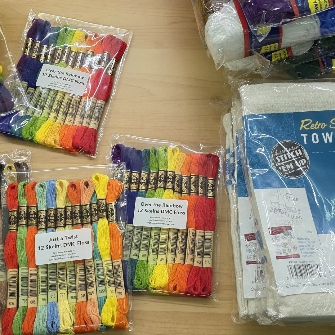 A wood table top stacked with craft supplies like packs of floss, yarn, and dish towels 