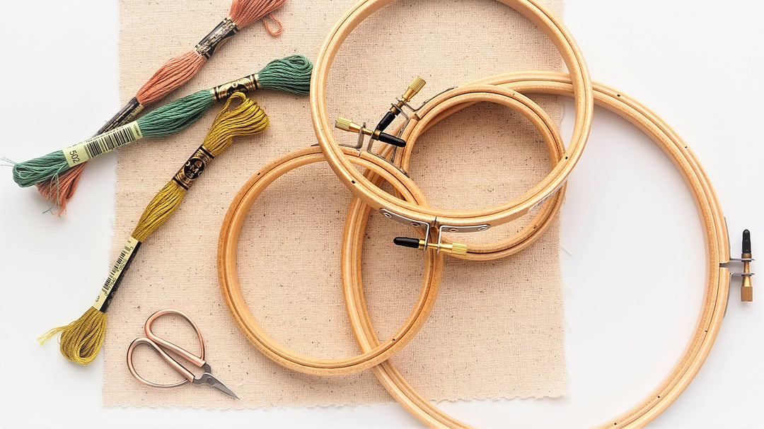An assembly of hand embroidery supplies, including a cotton fabric square, three skeins of floss, a small scissors, and four embroidery hoops in assorted sizes.