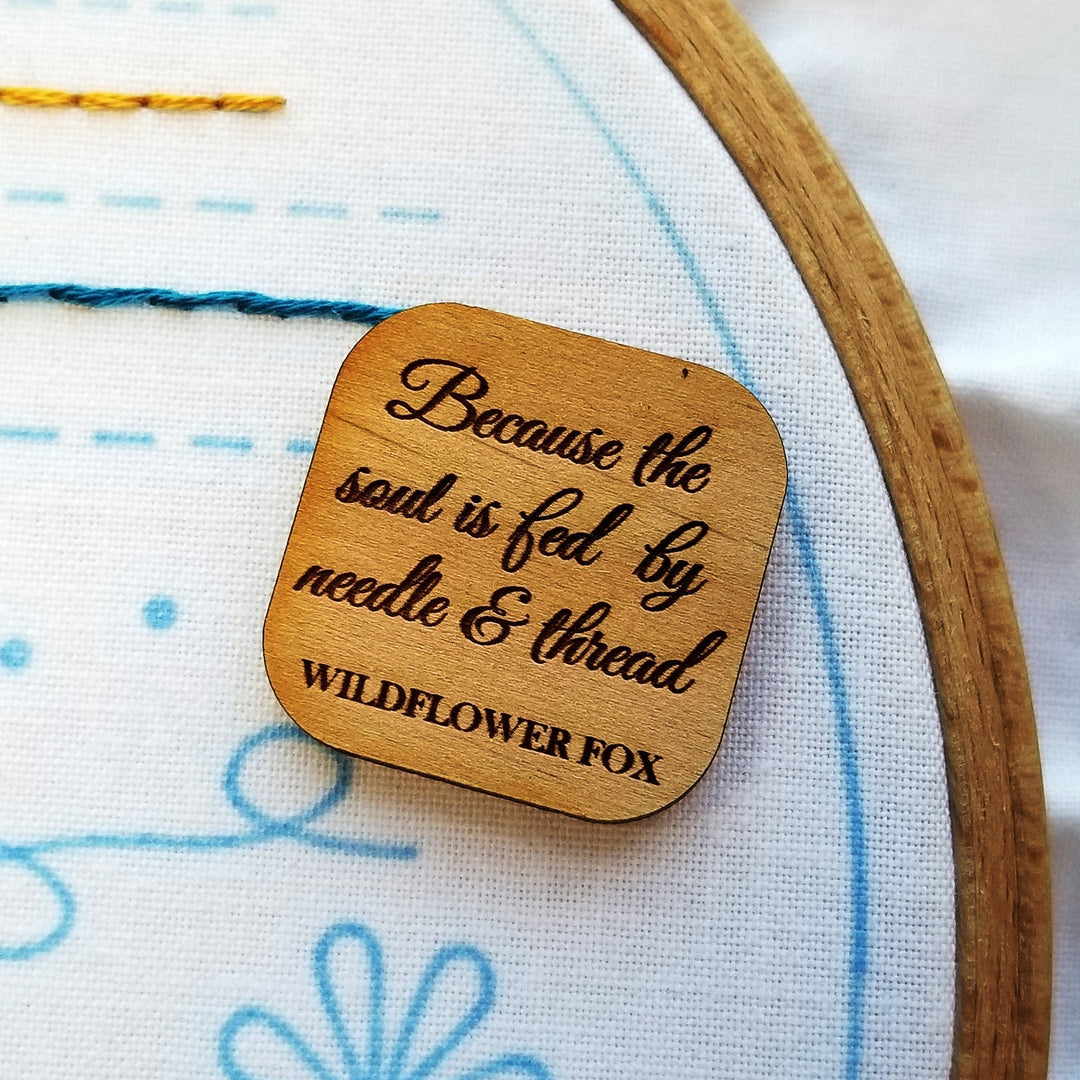 A small square wood needle minder laser engraved with the words "Because the soul is fed by needle and thread"