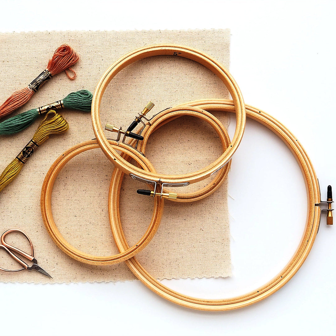 An assembly of hand embroidery supplies, including a cotton fabric square, three skeins of floss, a small scissors, and four embroidery hoops in assorted sizes.