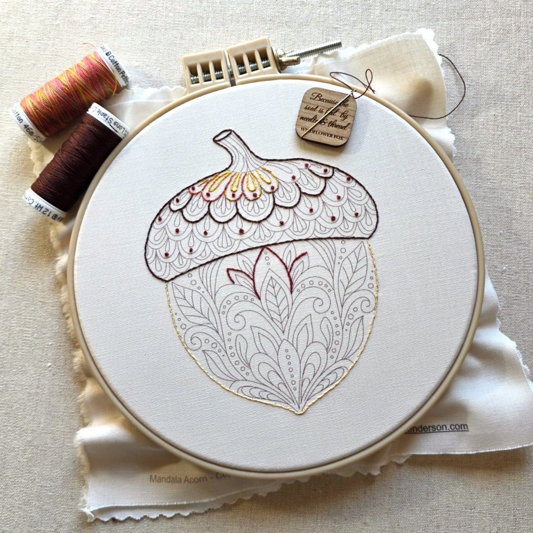 An embroidery hoop holding ivory fabric with a large acorn motif outlined in brown and filled with delicate floral patterns.  The hoop is styled on a beige fabric surface with thread spools and a wooden needle minder.