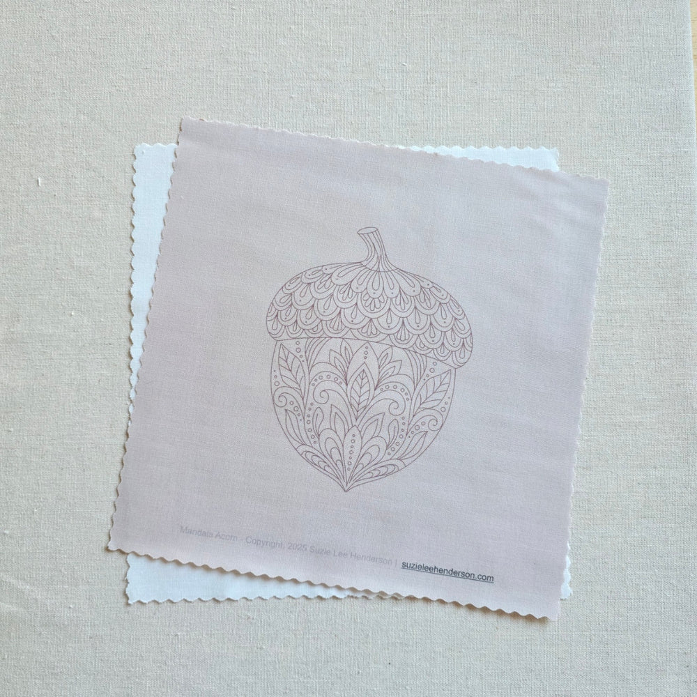 A square fabric printed with a delicate line-art illustration of an acorn with ornate leaf patterns. The fabric is layered over a white square of fabric on a textured beige linen background.