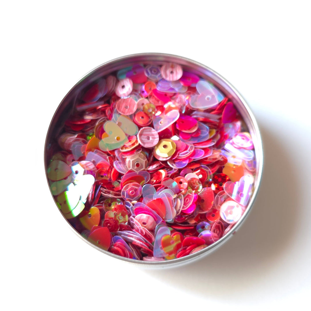A top-down view of a round metal tin filled with colorful decorative sequins and small embellishments on a white background.